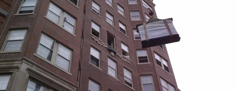 Wrapped piece being hoisted past the windows of a brick apartment building